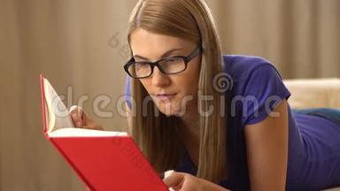 美丽迷人的年轻女人穿着紫罗兰色的t恤和一副黑色眼镜躺在沙发上看书。