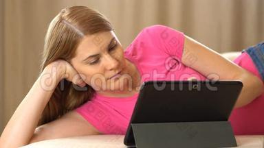 漂亮迷人的年轻女人穿着一件粉红色的t恤，一<strong>台</strong>平板电脑躺在沙发上。 浏览互联网和微笑
