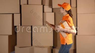 漂亮的女仓库工人穿着橙色制服走在棕色纸板箱周围。 4K视频