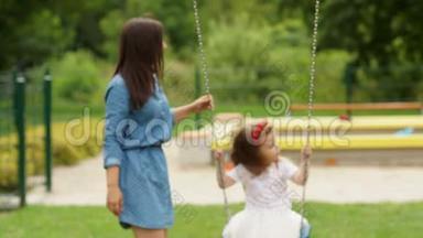 漂亮的布鲁内特和她可爱的女儿在游乐场玩耍的肖像。 小女孩骑着秋千出门