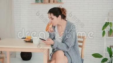 年轻女子在厨房里吃早餐，用智能手机浏览互联网。 女孩吃羊角面包，喝咖啡。