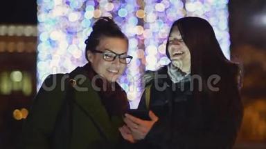 微笑姐妹使用手机在一条带有圣诞灯装饰的夜街。 两个布鲁内特女孩笑