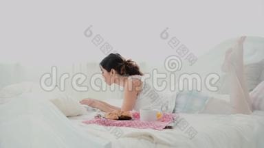 年轻漂亮的女人躺在床上用笔记本电脑。 女孩早<strong>上上</strong>网，吃早餐