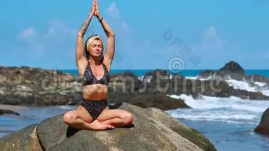 美丽的金发女孩穿着黑色上衣，在瑜伽中放松，在紫色的垫子上摆莲花，对抗海浪在岩石上奔跑