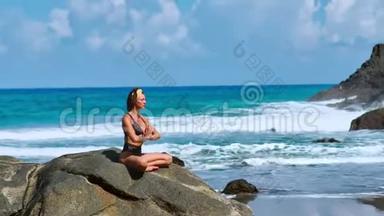 美丽的金发女孩穿着黑色上衣，在瑜伽中放松，在紫色的垫子上摆莲花，对抗海浪在岩石上奔跑