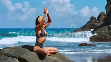 美丽的金发女孩穿着黑色上衣，在<strong>瑜伽</strong>中放松，在<strong>紫色</strong>的垫子上摆莲花，对抗海浪在岩石上奔跑