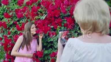 年长的女人拍照年轻的女孩。 妈妈给女儿拍照。 红玫瑰花在背景上。