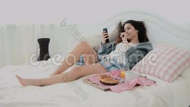 年轻微笑的女人躺在床上。 布鲁内<strong>特</strong>女孩在早餐期间上网冲浪，使用智能手机和<strong>饮</strong>料<strong>果汁</strong>。