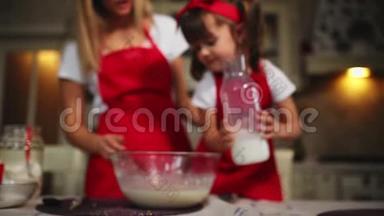 漂亮的年轻母亲帮她的小女儿用红色围裙做蛋糕。<strong>倒牛奶</strong>搅拌面糊