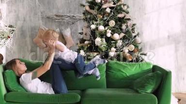 妈妈带着一个孩子在圣诞树背景的沙发上玩耍。 圣诞节室内