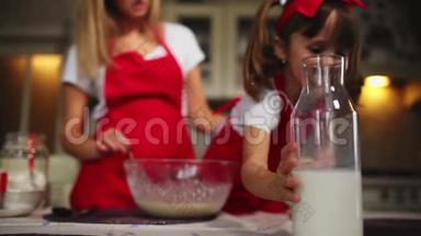 漂亮的年轻母亲帮她的小女儿用红色围裙做蛋糕。倒牛奶搅拌面糊