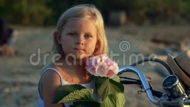 那个金发女孩坐在<strong>海边</strong>的沙滩上，手里拿着一朵粉<strong>红色</strong>的花。 美丽的女孩