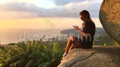 漂亮的女人坐在悬崖边，用智能手机发短信。 泰国普吉岛。 高清慢动作惊人的视野。