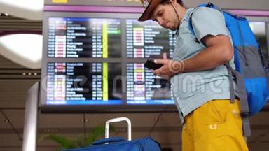 年轻英俊的国际机场游客戴着帽子，用手机站着，背包和手提箱在