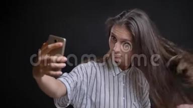 年轻的白种人长发女人在<strong>黑色背景</strong>下使用智能手机制作自拍<strong>照片</strong>的肖像。