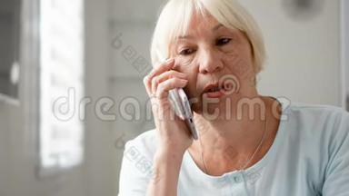 年长的女人坐在家里使用智能手机。 离退休妇女用手机说话