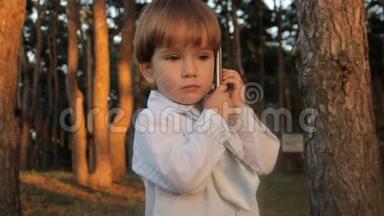 一个穿着黑色马裤的小男孩，一件带领带的白色衬衫在智能手机上交谈。 夏天日落。
