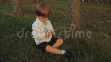 一个穿着黑色马裤的小男孩，一件带领带的白色衬衫，坐在绿草上，玩着智能手机。