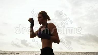 运动的年轻女子在海上背着太阳打拳击。 美丽的女拳击手在海滩上训练