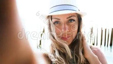 美丽的金发女郎戴着帽子，在日落时分在沙滩上自拍