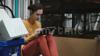 年轻女子坐在仓库里，用平板电脑检查货物清单。 有手推车的人走到摄像机前面。 4K