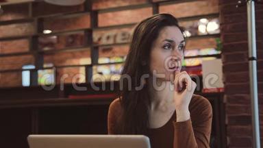 年轻的黑发女孩在咖啡馆里用笔记本电脑工作，沉思地看着窗外。
