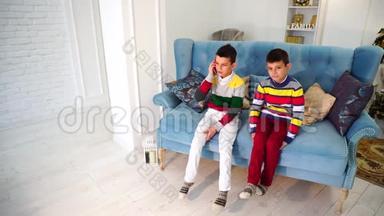 两个男孩<strong>弟弟</strong>轮流和奶奶在手机上交谈，白天坐在明亮的房间里的蓝色沙发上。