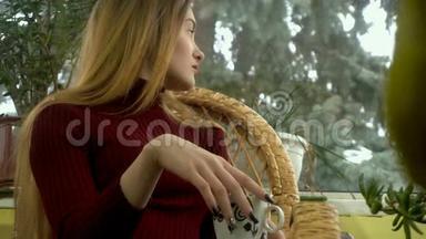 年轻女子坐在一把柳条椅子上，望着窗外，身后降着雪，享受着一杯