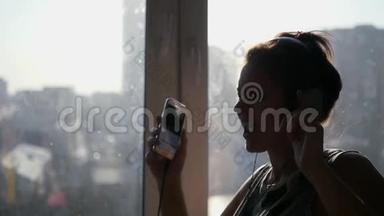 一个年轻开朗的女孩从电话里用耳机听音乐，在阳光下在窗户边大声唱歌跳舞