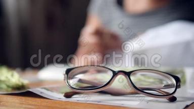 老年妇女躺在桌子上的眼镜和妇女在Blury背景下做十字绣霍比。 4K，慢动作。