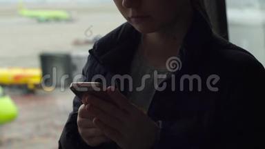 一名年轻女子<strong>在机场</strong>一扇大窗户前使用智能手机的特写镜头