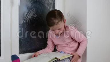 小女孩坐在冰冻的窗户后面的窗台上。 可爱的小女孩坐在窗台上读日记。