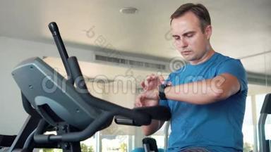 一个英俊健康的年轻人在健身房骑着健身车做运动。用他的智能手表检查生命体征