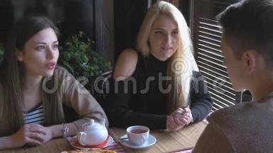 两个<strong>女孩</strong>和一个年轻人坐在一张桌子旁的咖啡馆里说话。 他们<strong>喝茶</strong>吃披萨。 他们告诉新闻，他们