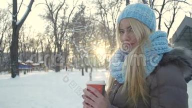 迷人的年轻女人拿着一杯咖啡，在冬天公园的背景上<strong>暖手</strong>。