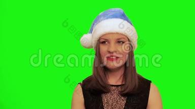戴着圣诞帽的迷人女人笑着，唱着圣诞歌，对着彩色的绿色屏幕跳舞