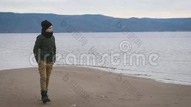 穿夹克，戴黑色帽子，脖子上戴围巾的男孩在海滩上散步。 <strong>背景</strong>下的一种观点.. 美丽的<strong>大海</strong>