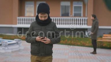 穿着<strong>暖色</strong>灰色夹克，戴着黑色帽子和围巾的男孩正在街上用电话站着，在他身后