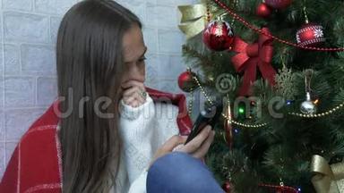 一个女人坐在一个有红色格子的地板上，在圣诞树旁边使用智能手机