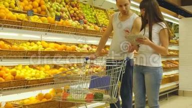 年轻的白种人夫妇带着市场手推车在超市里散步，选择新鲜的<strong>苹果</strong>。 男人在放水果