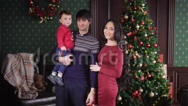 爸爸妈妈和他们的小儿子一起看着镜头，背景上穿着圣诞树。 那个男孩
