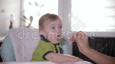 好奇可爱的宝宝环顾四周，坐在婴儿椅上。 妈妈用勺子喂她可爱的一岁小孩。
