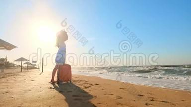 一个年轻的女人带着一个手提箱坐在海滩上。 一个年轻的女孩带着一个轮式手提箱沿着海滩散步。 一路走来