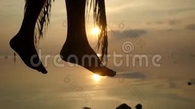 在美丽的夕阳下，在热带海滩挥舞着雄腿的剪影。 年轻人<strong>坐在</strong>棕榈<strong>树上</strong>。 慢动作