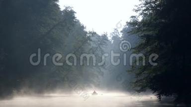 孤独的人，带着桨在船上，在黎明的雾中平静的河流上游泳。 清晨的第一缕阳光使他们