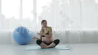 美丽的怀孕少女大肚腩正在健身房里跳舞坐在地板上莲花的位置..