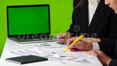 办公室里的办公桌上，男人和女人画一张图表，打开带有绿色屏幕的笔记本
