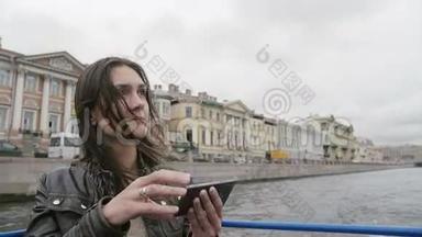 年轻女子在河边旅游，拍照，使用智能手机。 风吹头发。 圣彼得堡的城市景色.. 慢节奏
