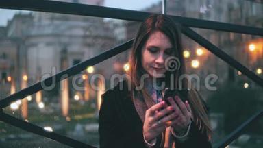 年轻漂亮的女人晚上坐在城市里使用智能手机。 女孩用触摸屏键盘发短信。