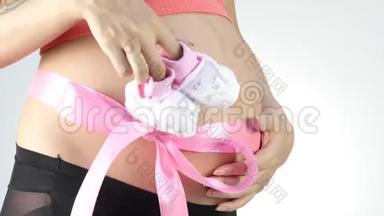 穿着内衣的怀<strong>孕妈妈</strong>在玩婴儿鞋。 腹部有粉红色的丝带。 慢动作